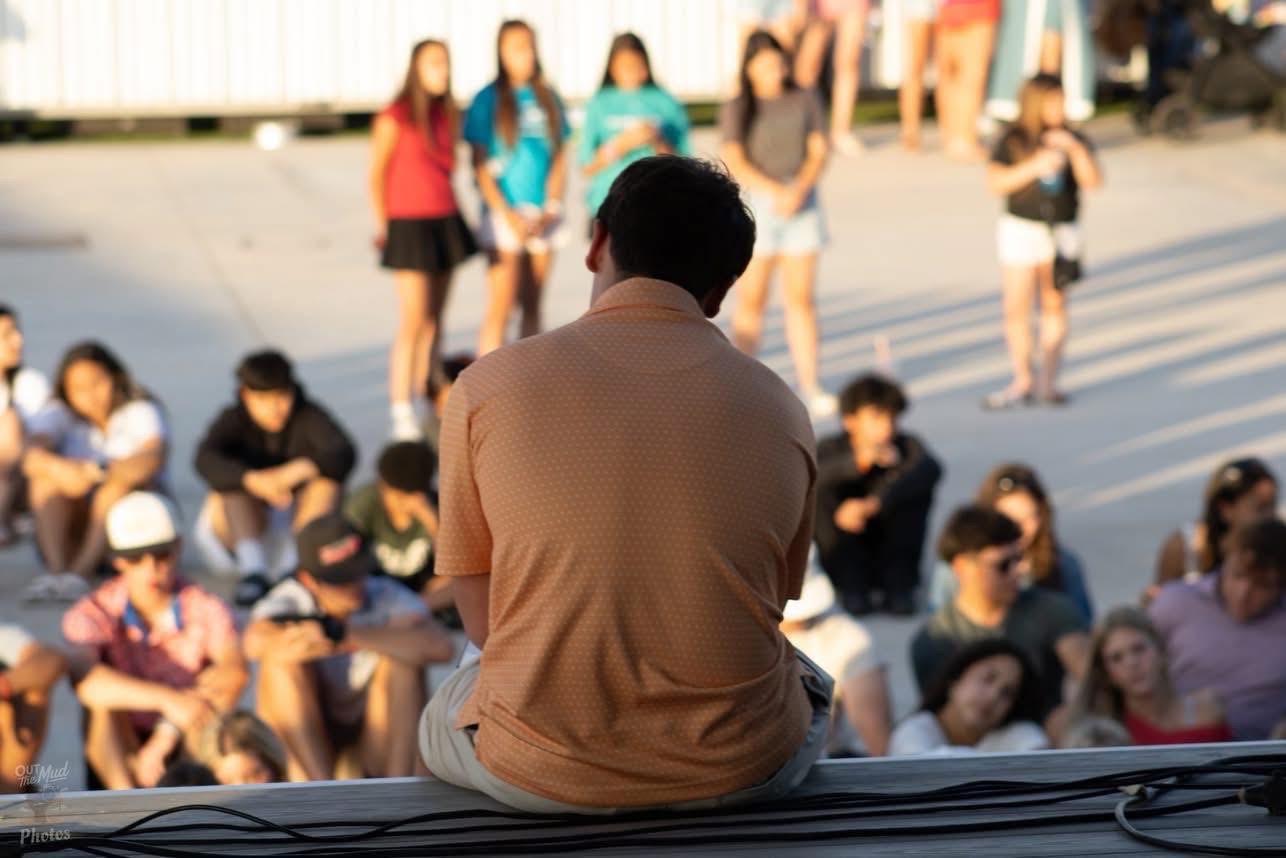Tucker seated on the edge of a stage, speaking with an outdoor audience.